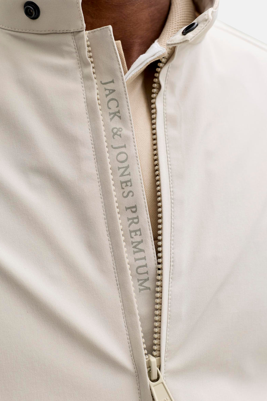 Beige korte jas van PREMIUM by JACK & JONES, met ritssluiting, verticale JACK & JONES PREMIUM print en zwarte drukknoop, over een lichtbruin poloshirt.