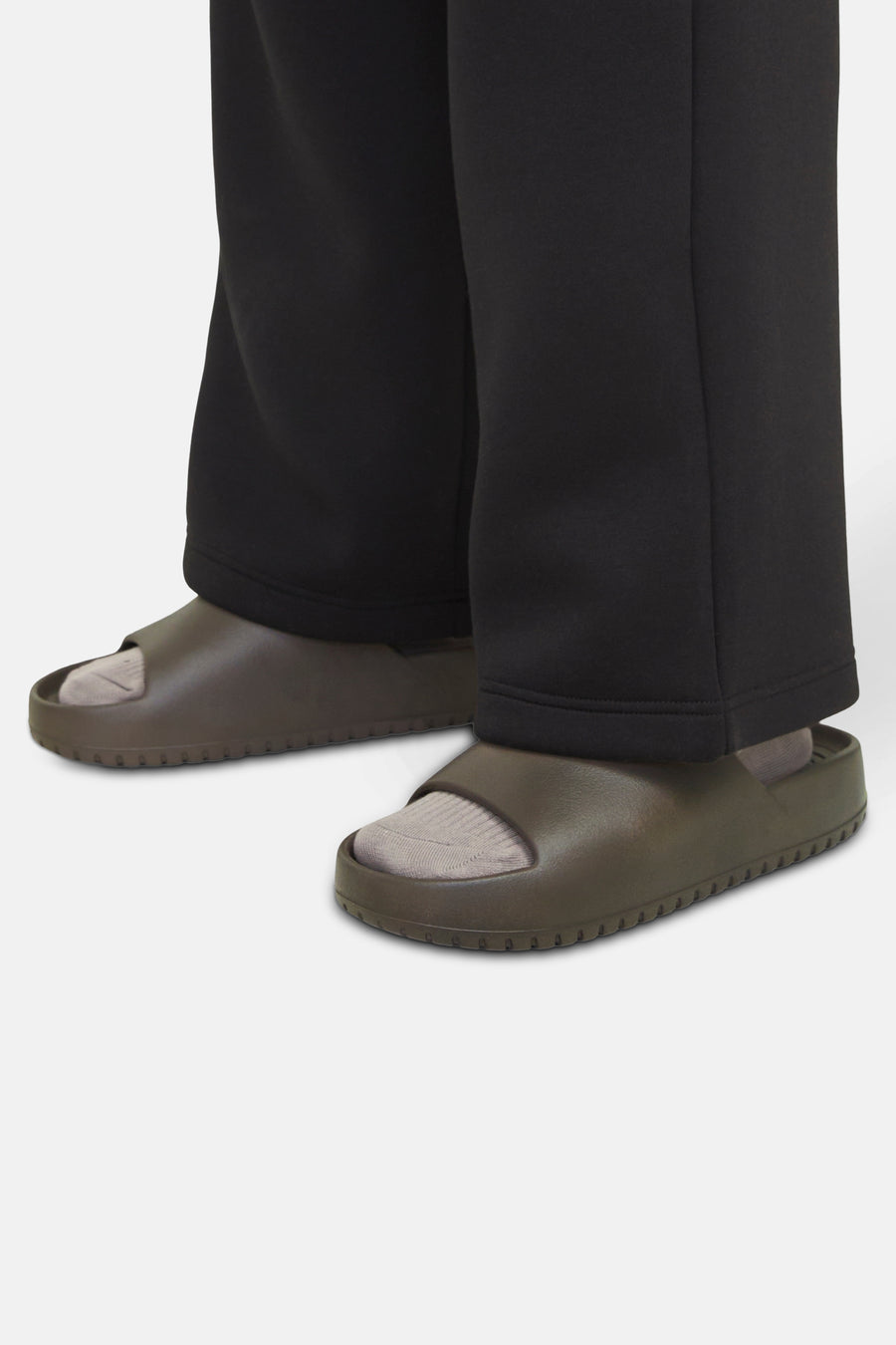 Persoon met zwarte joggingbroek en kaki sandalen met sokken, staand op een witte achtergrond.