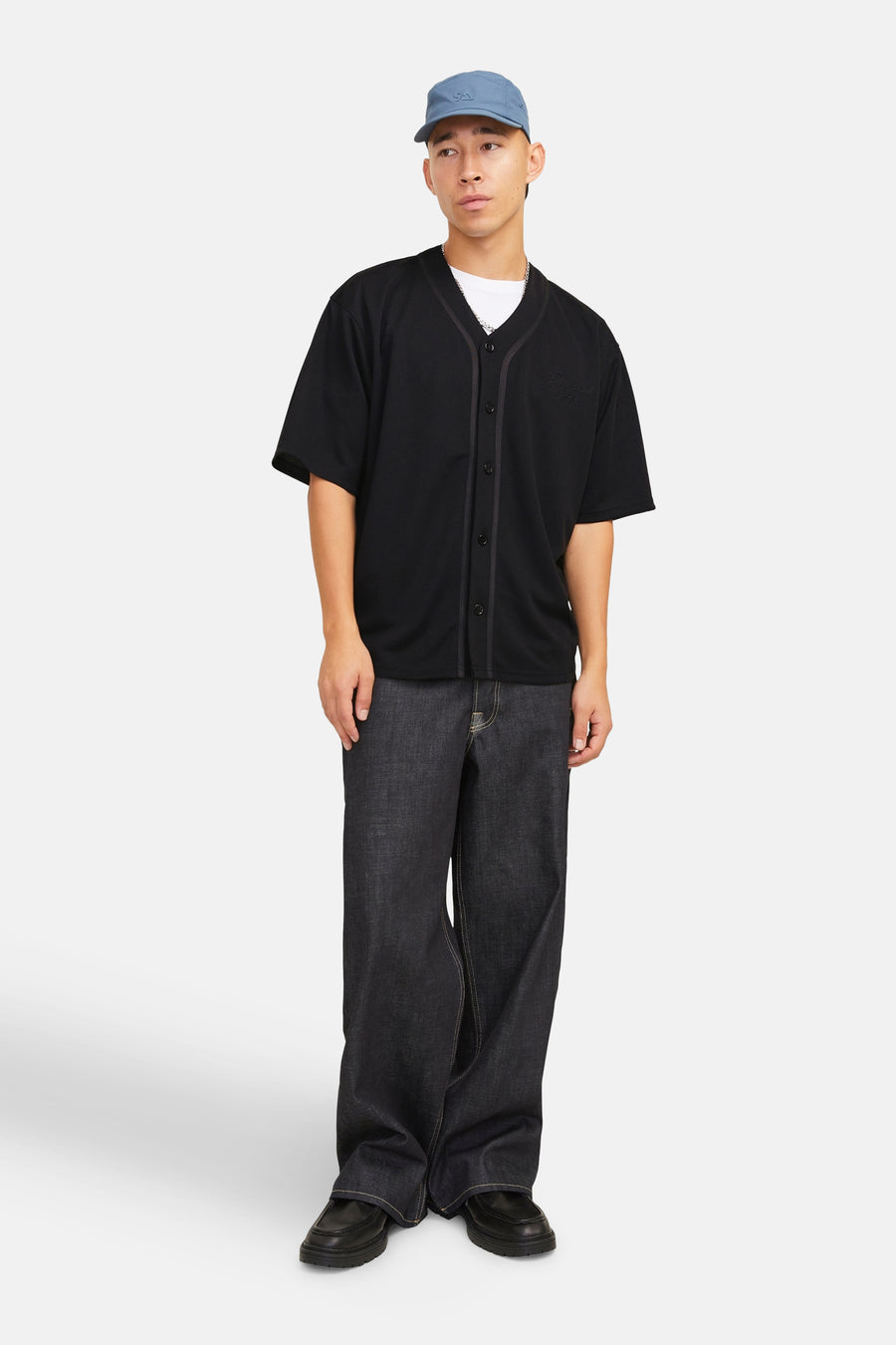 Man met blauwe pet, zwart baseballshirt, losse jeans en zwarte schoenen, staand voor een witte achtergrond.