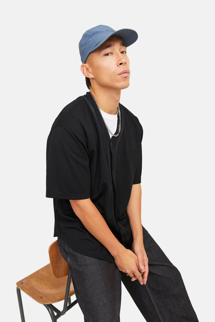 Man met blauwe pet, zwarte blouse en donkere jeans zittend op een houten stoel.