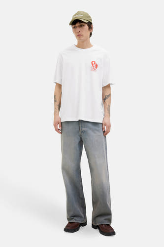 Wit T-shirt met korte mouwen van ORIGINALS BY JACK & JONES, met een rode print op de borst, gedragen met een pet, wijde jeans en bruine loafers.