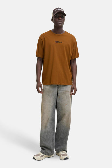 T-shirt brun à manches courtes d'ORIGINALS BY JACK & JONES, avec l'inscription Kyoto, porté avec un chapeau et des jeans larges.