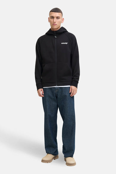 Gilet noir de CORE BY JACK & JONES, avec fermeture éclair et poches à l'avant, associé à un T-shirt blanc, des jeans décontractés et des chaussures beige.