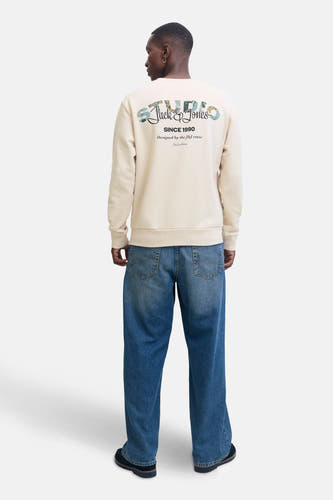 Gilet beige de CORE BY JACK & JONES, avec logo sur le dos, associé à des jeans larges et des chaussures noires.