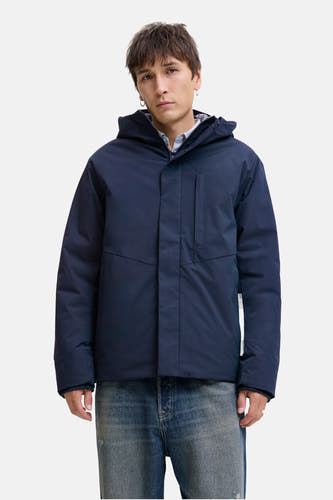 Blauwe, korte jas van CORE BY JACK & JONES, met capuchon, gedragen over een gestreept overhemd en een blauwe jeans.

