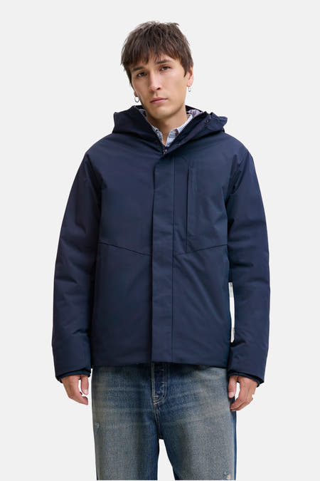 Blauwe, korte jas van CORE BY JACK & JONES, met capuchon, gedragen over een gestreept overhemd en een blauwe jeans.
