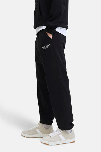 Zwarte joggingbroek van JACK & JONES JEANS INTELLIGENCE, met witte tekst op het been, gecombineerd met crèmekleurige sneakers en een hoodie.
