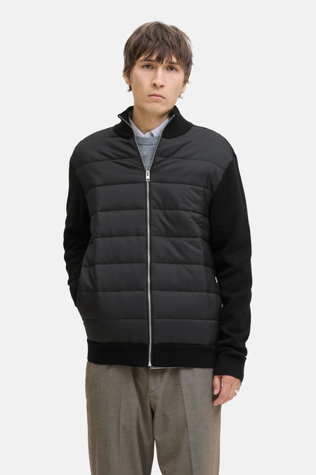 Man met zwarte jas met gewatteerd voorpand, grijze trui en bruine broek, staand.