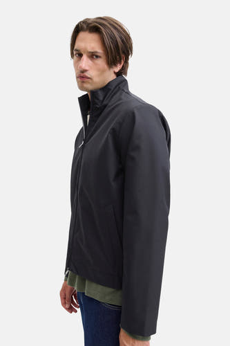 Manteau court noir de JJREBEL, vu sur un homme portant une chemise verte olive et des jeans noirs.