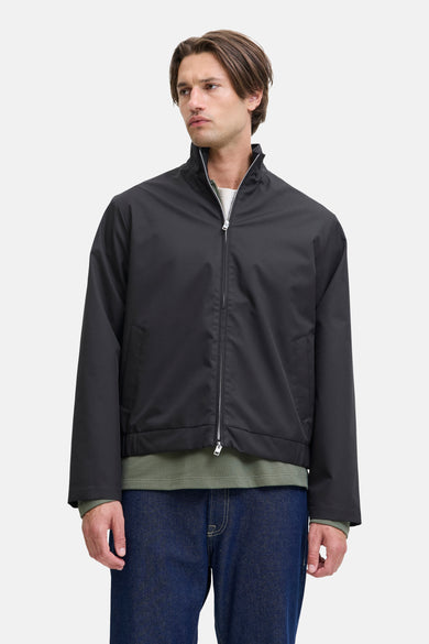 Zwarte korte jas van JJREBEL, met lange mouwen en zilverkleurige ritssluiting, gedragen over een olijfgroen shirt en donkerblauwe jeans.
