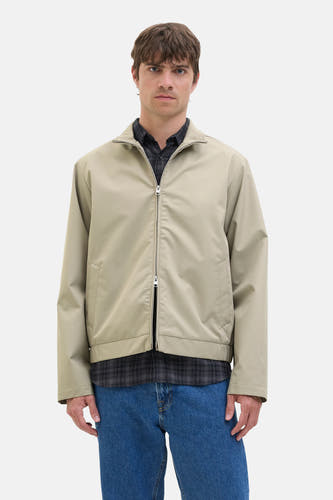 Manteau court couleur écru de JJREBEL, avec fermeture éclair et porté sur un chemisier à carreaux et des jeans bleus.