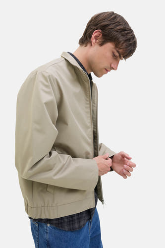 Manteau court écru de JJREBEL, avec fermeture éclair, associé à une chemise à carreaux et un pantalon en denim.