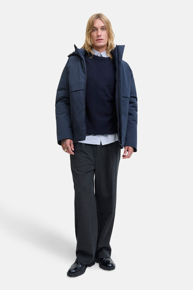 Blauwe trui met ronde hals van PREMIUM by JACK & JONES, gedragen over een gestreept overhemd, met grijze pantalon en zwarte schoenen.
