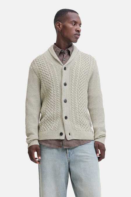 Beige gilet van PREMIUM BLUE by JACK & JONES, met kabelpatroon en donkere knopen, gedragen over een bruin shirt en lichtblauwe jeans.
