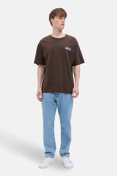 Bruin T-shirt met korte mouwen van JJREBEL, met een lichtblauwe print linksboven, gedragen met een lichte jeans en witte sneakers.