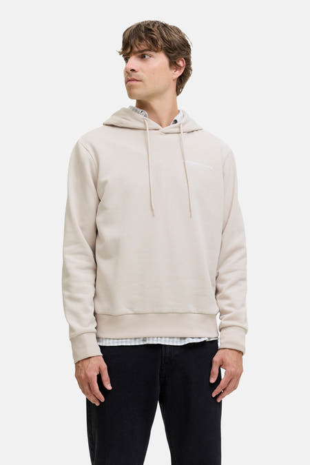 Ecru sweater met kap van JJREBEL, gedragen over een geruit overhemd en met zwarte jeans.
