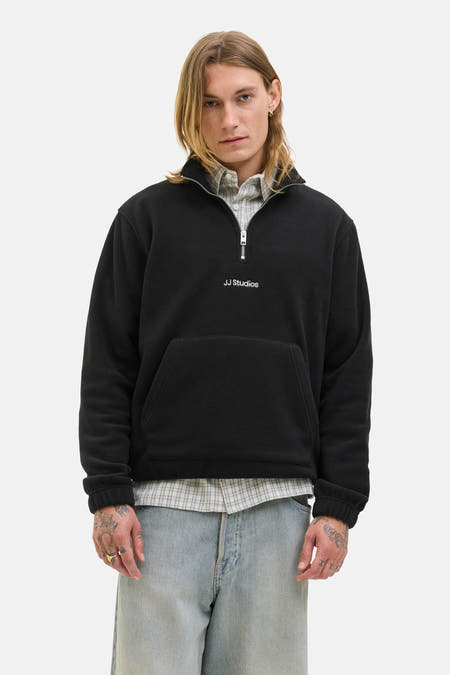 Fleece noir de JJ Studios, avec une fermeture éclair à moitié et une poche poitrine, porté sur un chemisier rayé et un pantalon en denim.