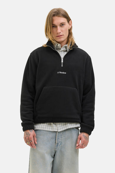Fleece noir de JJ Studios, avec une fermeture éclair à moitié et une poche poitrine, porté sur un chemisier rayé et un pantalon en denim.