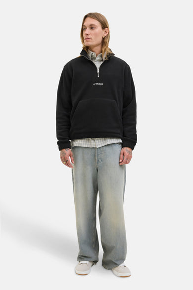 Fleece noir de CORE BY JACK & JONES, avec une fermeture à glissière partielle, une poche et porté sur un chemisier rayé avec des jeans larges.