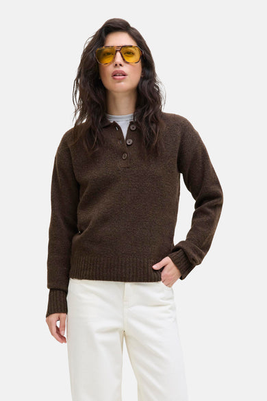 Pull-over marron de JJXX, avec fermeture à boutons, porté sur un chemise grise et associé à un pantalon blanc et des lunettes de soleil jaunes.