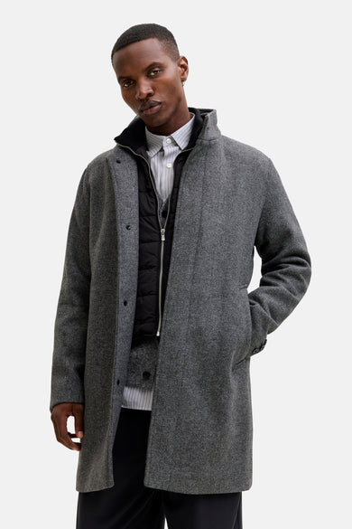 Lange grijze jas van PREMIUM by JACK & JONES, met visgraatmotief, gedragen over een gewatteerd vest en gestreept overhemd.