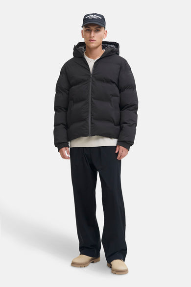 Zwarte korte jas van CORE BY JACK & JONES, met capuchon, gecombineerd met een witte trui, zwarte broek en beige schoenen.