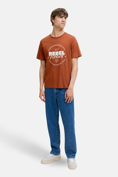 T-shirt marron à manches courtes de JJREBEL, avec une impression blanche sur la poitrine, associé à un jeans bleu et des baskets.