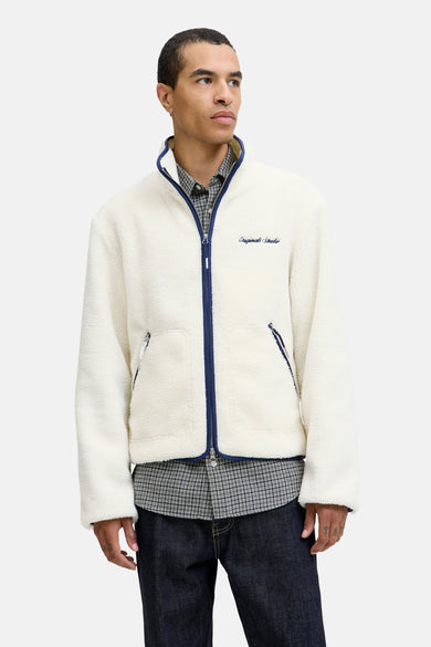 Manteau beige d'ORIGINALS BY JACK & JONES, avec des fermetures éclair bleues et des poches, porté sur un chemisier à carreaux et des jeans foncés.