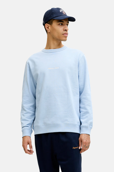 Blauwe sweater van ORIGINALS BY JACK & JONES met lichte 'Originals Studio' tekst op de borst, gedragen door een man met donkerblauwe pet en joggingbroek.