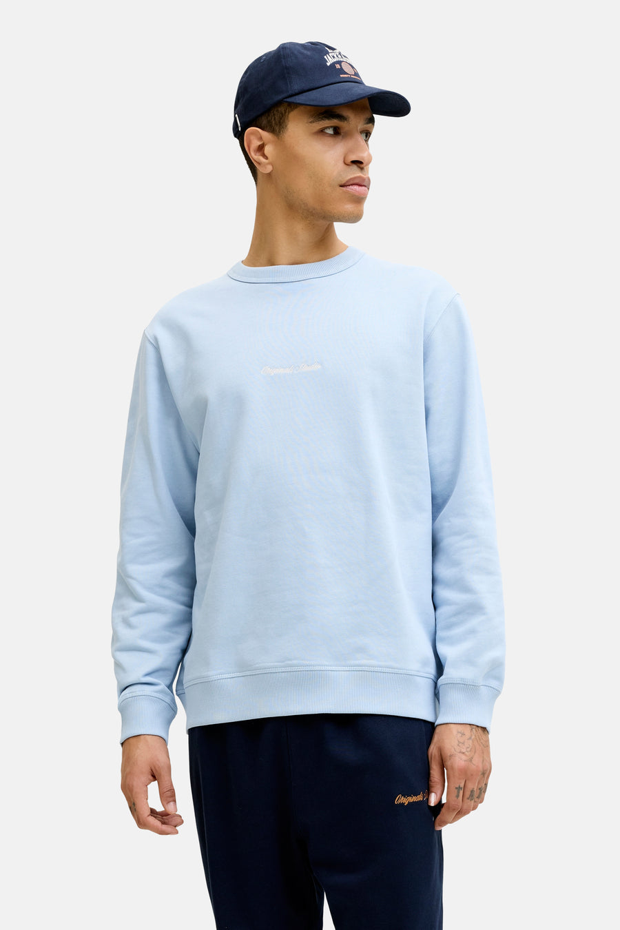 Blauwe sweater van ORIGINALS BY JACK & JONES met lichte 'Originals Studio' tekst op de borst, gedragen door een man met donkerblauwe pet en joggingbroek.