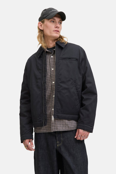 Zwarte korte jas van ORIGINALS BY JACK & JONES, gedragen over een geruit overhemd met donkere jeans en een pet.
