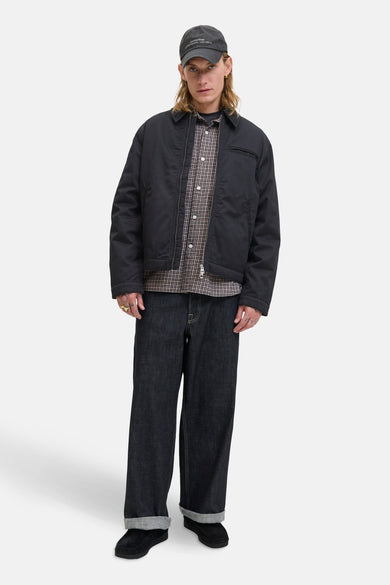 Zwarte korte jas van ORIGINALS BY JACK & JONES, gedragen over een geruit overhemd en een donkere jeans, met een pet.
