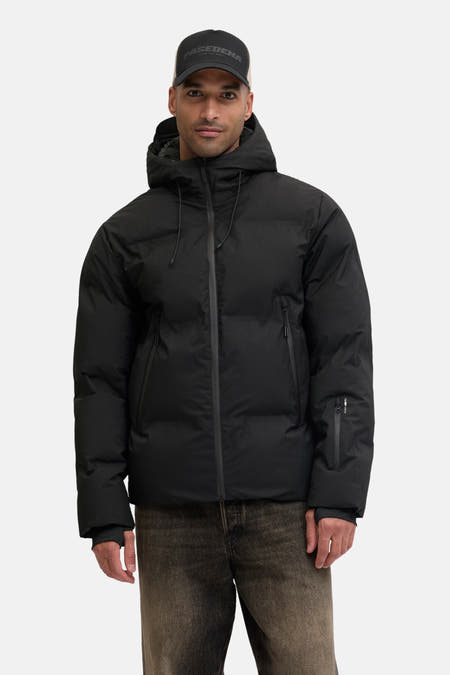 Zwarte, korte jas van CORE BY JACK & JONES, met capuchon en rits, gecombineerd met een pet en jeans.