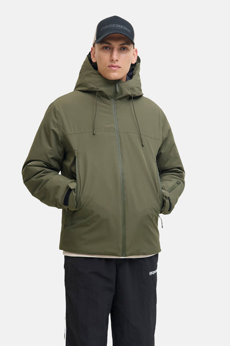 Veste courte verte de CORE BY JACK & JONES avec capuche, portée par un homme avec une casquette noire, un pull de couleur crème, un jogging noir et des baskets beiges.