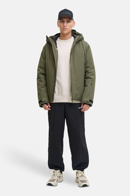 Groene korte jas van CORE BY JACK & JONES met capuchon, gedragen door een man met een zwarte pet, crèmekleurige trui, zwarte joggingbroek en beige sneakers.