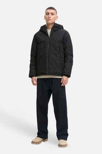 Zwarte korte jas van CORE BY JACK & JONES met capuchon, rits en zakken, gecombineerd met een lichtbeige trui, donkerblauwe broek en beige schoenen.