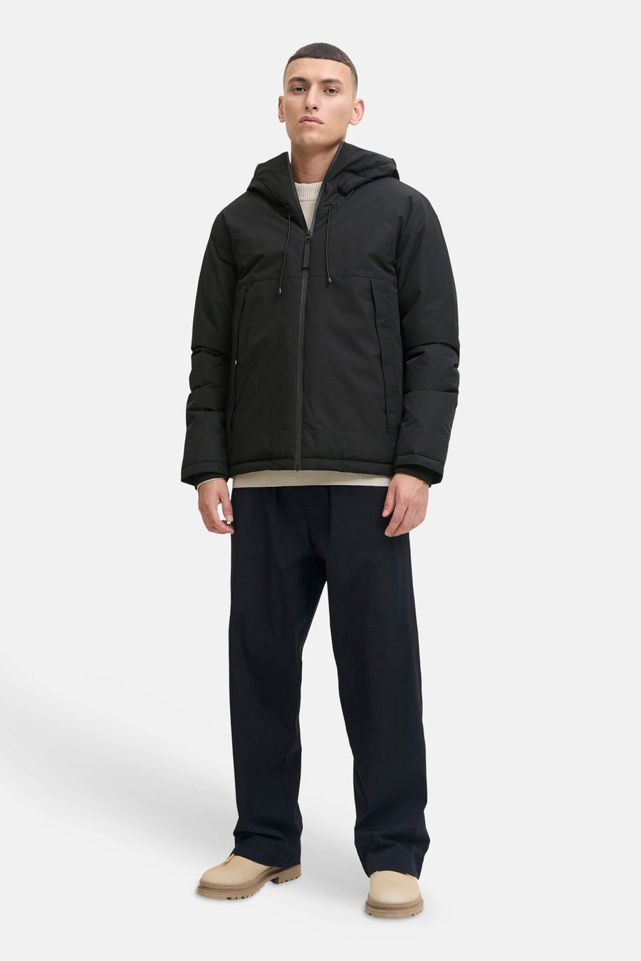 Zwarte korte jas van CORE BY JACK & JONES met capuchon, rits en zakken, gecombineerd met een lichtbeige trui, donkerblauwe broek en beige schoenen.