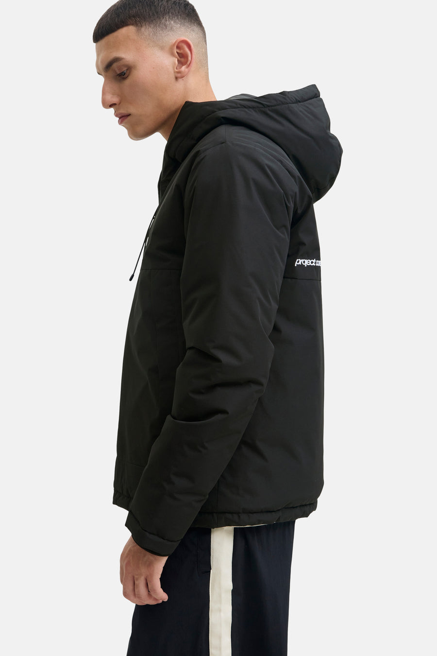 Zwarte korte jas van CORE BY JACK & JONES met capuchon en wit logo op de mouw, gedragen door een man in zijzicht.