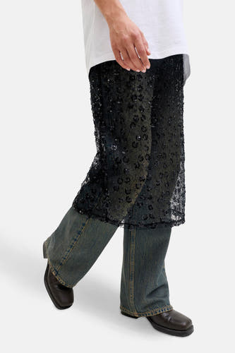 Lange zwarte rok van JJXX, met een doorschijnende stof met bloemmotief en pailletten, over een blauwe jeans gedragen.
