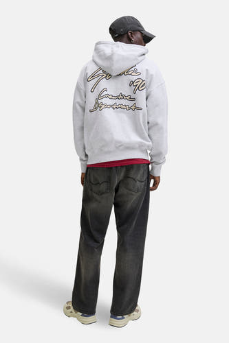 Pull-over gris avec capuche d'ORIGINALS BY JACK & JONES, avec une impression noire et dorée sur le dos, porté avec un chapeau gris foncé et des jeans sombres.
