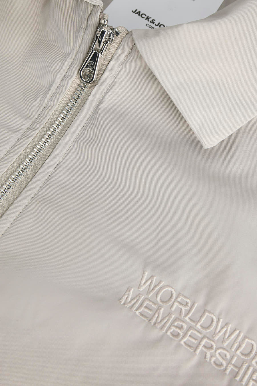 Grijze korte jas van CORE BY JACK & JONES, met zilverkleurige ritssluiting en 'Worldwide Membership'-borduring.