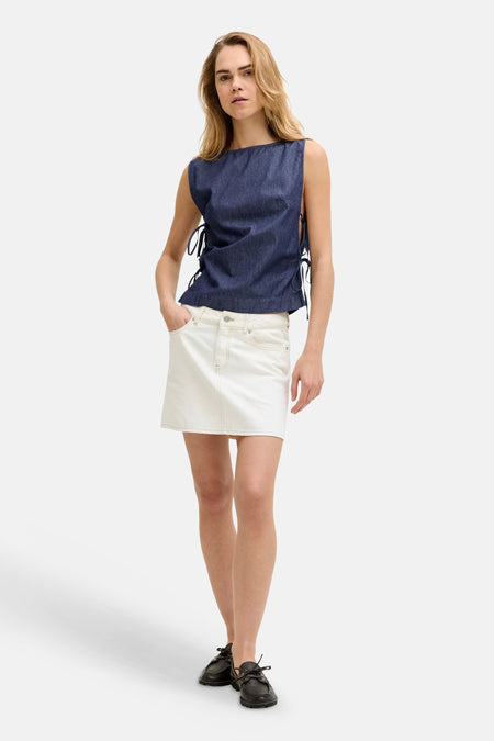 Donkerblauwe denim mouwloze blouse van JJXX met strikjes aan de zijkanten, gedragen met een witte rok en donkere instappers.
