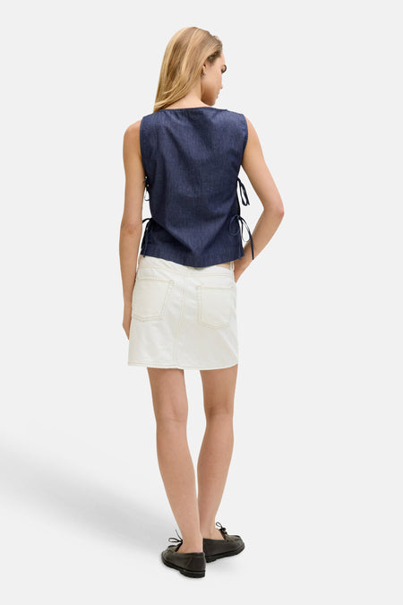 Donkerblauwe mouwloze denim blouse van JJXX met strikdetails aan de zijkanten, achteraanzicht, gecombineerd met een witte spijkerrok.