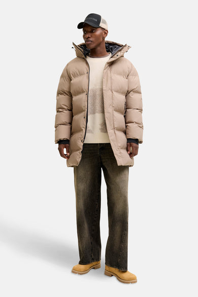 Beige trui met ronde hals van CORE BY JACK & JONES, gedragen onder een kaki pufferjas met capuchon, gecombineerd met donkere jeans en bruine veterschoenen.