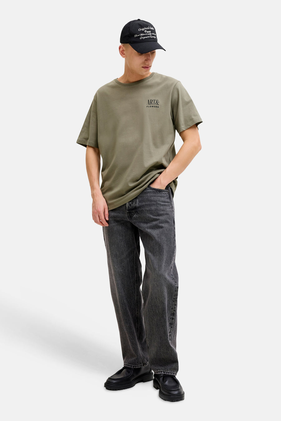 T-shirt de couleur kaki de ORIGINALS BY JACK & JONES, à manches courtes et avec un petit imprimé, porté avec une casquette noire et un jean gris.