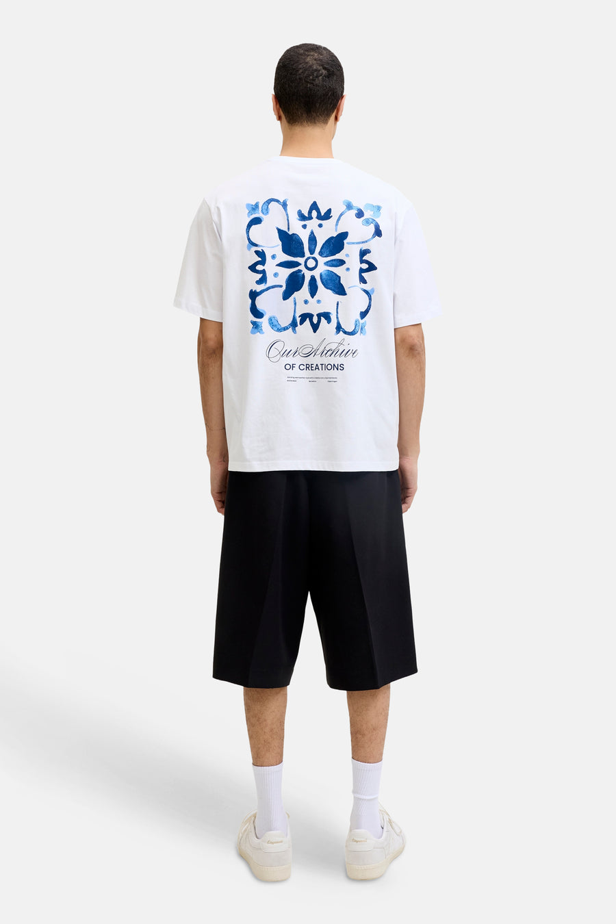 Wit T-shirt van ORIGINALS BY JACK & JONES met een blauw floraal tegelpatroon en tekst op de rug, gedragen met zwarte korte broek en witte sneakers.