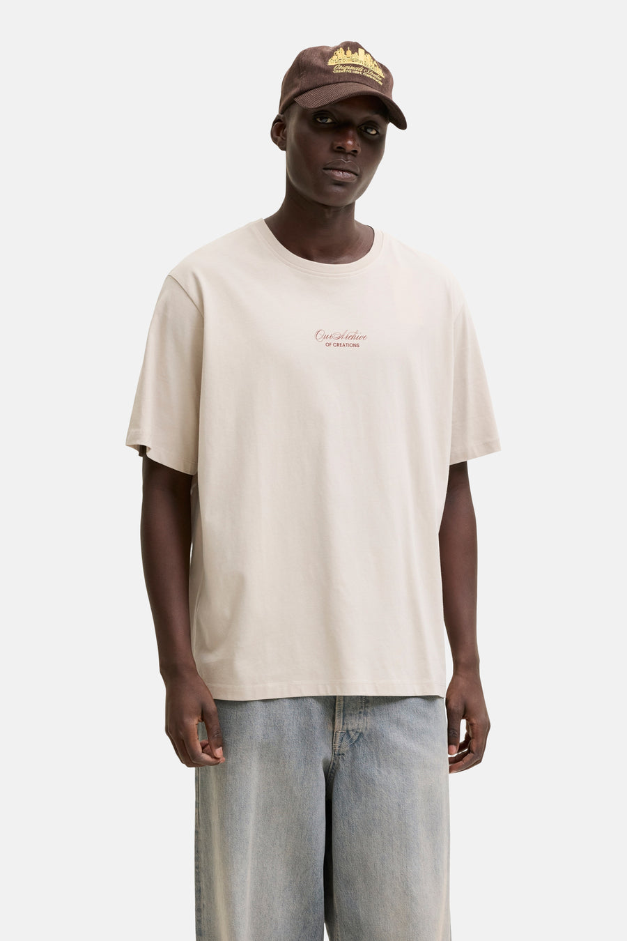 Beige T-shirt van ORIGINALS BY JACK & JONES met borstprint, gedragen door een man met jeans en een bruine pet.