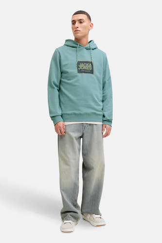 Groene sweater met kap van CORE by JACK & JONES, met een logo op de borst, gecombineerd met een blauwe jeans en witte schoenen.