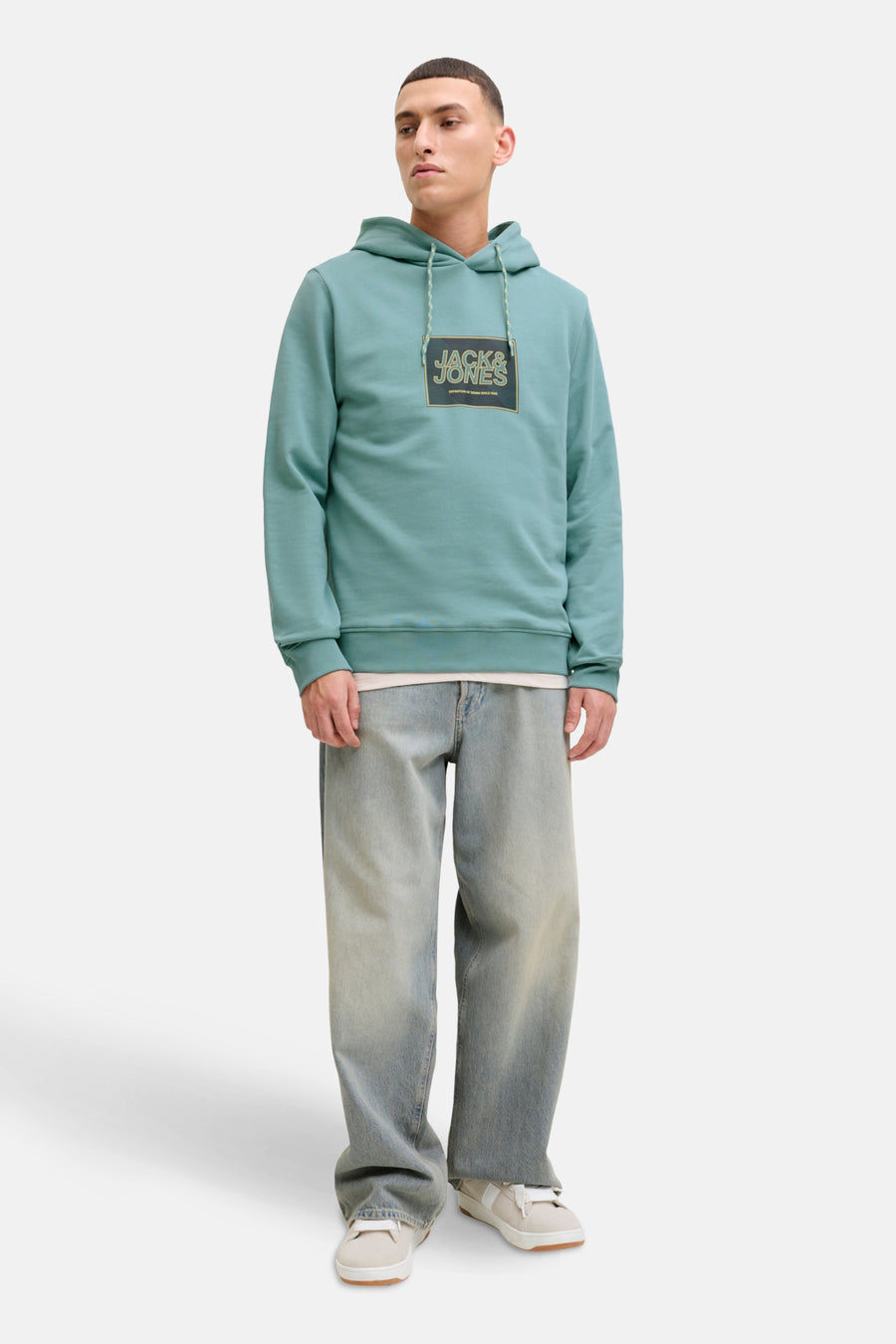 Groene sweater met kap van CORE by JACK & JONES, met een logo op de borst, gecombineerd met een blauwe jeans en witte schoenen.