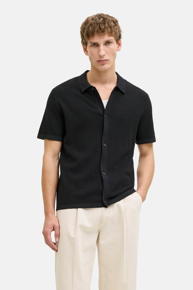 Pull noir de CORE BY JACK & JONES, à manches courtes et fermeture boutonnée, porté avec un débardeur blanc et un pantalon beige.